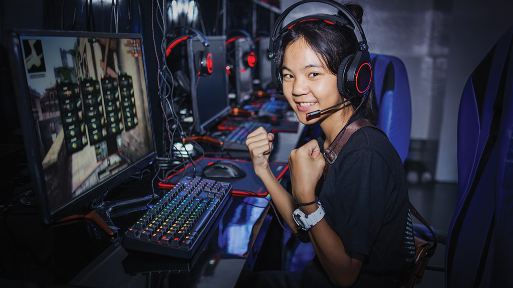 A girl in front of a computer wearing a headset gives a thumbs up to the camera