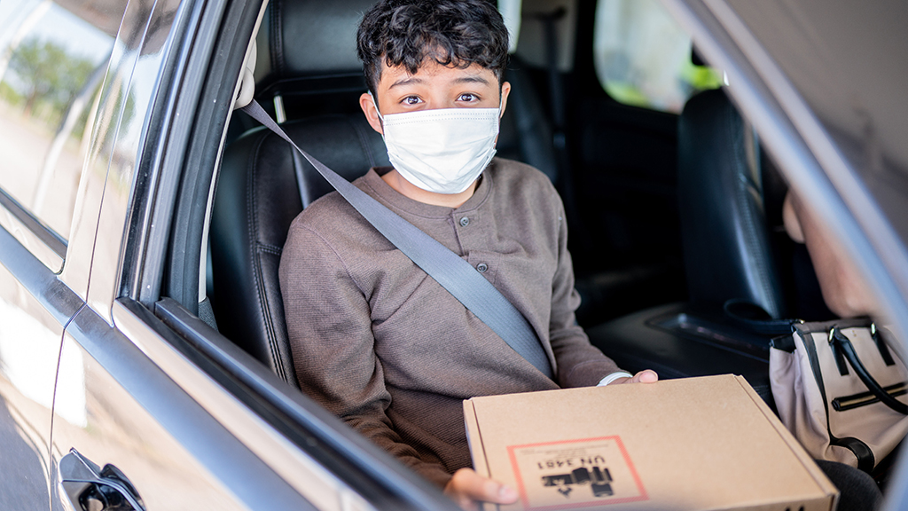 a student with a face mask in a car holds his new remote learning equipment 