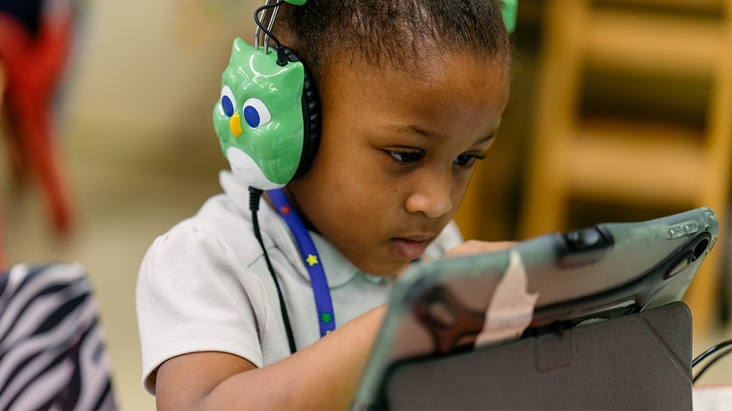 a child wearing owl headphones uses a digital device as part of a literacy program
