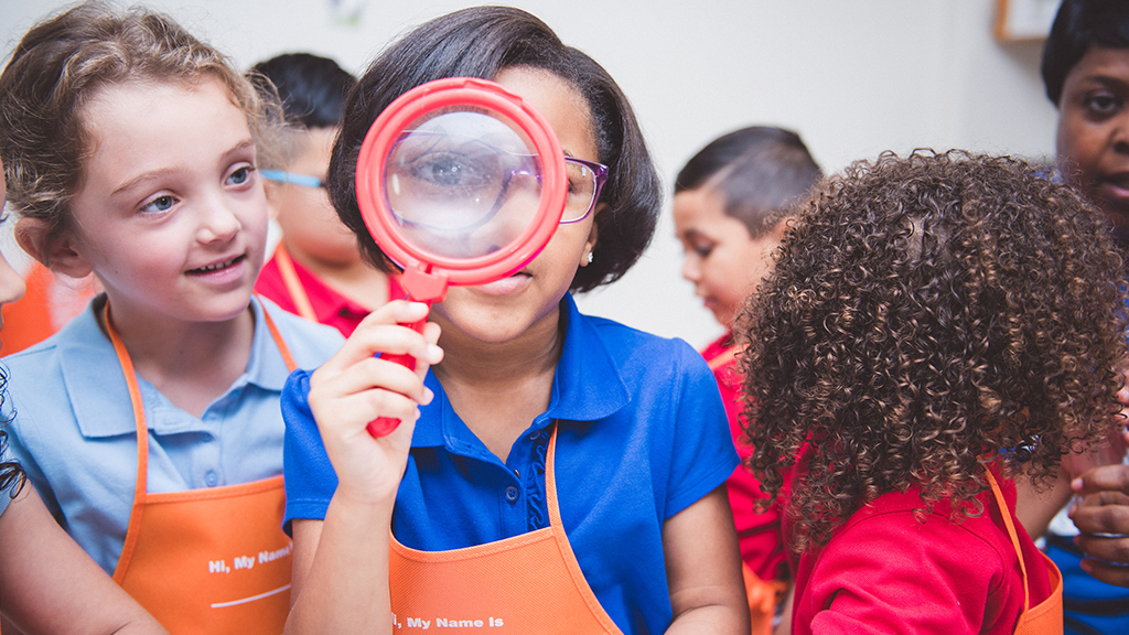 a student, surrounded by her classmates, looks through a magnifying glass
