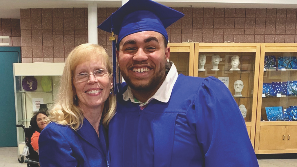 the superintendent of the district embraces a student on graduation day, they are both in academic regalia and the student is wearing a graduation cap