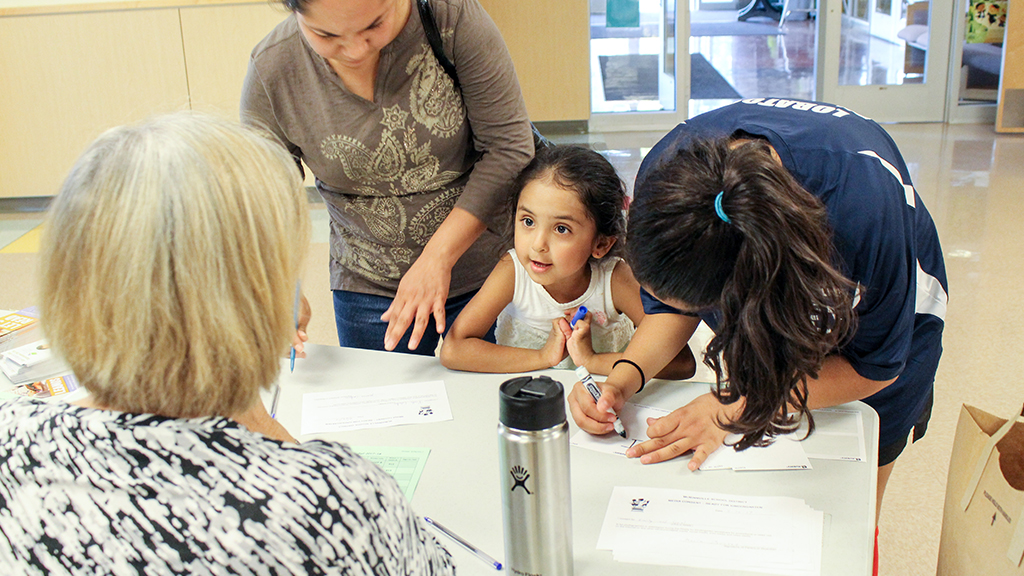 two adults check in at a table with a volunteer. the two adults are joined by a young girl, who is looking up at the volunteer