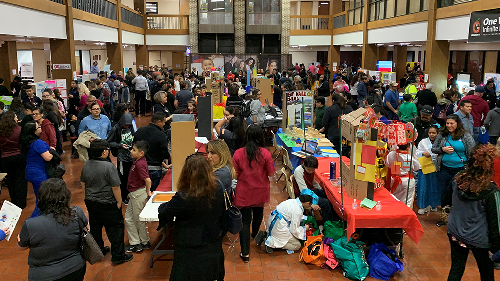 a crowd of people walk through a large room that has been set up to display projects at each table