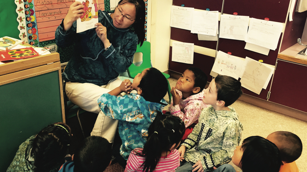 A teacher in a school in Alaska sits in a chair and reads a book to young children sitting on the floor. 
