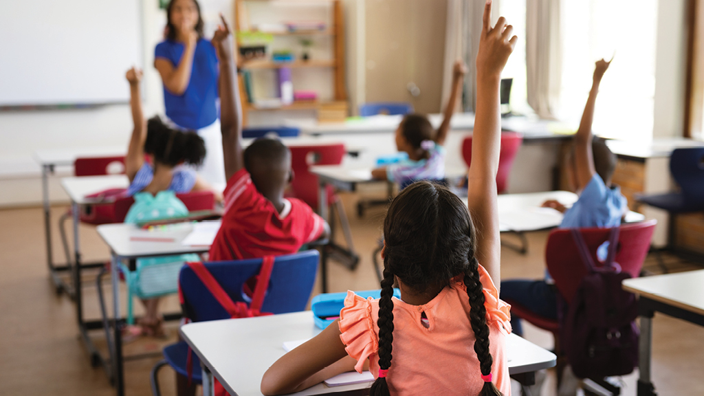 A teacher looks over a classroom of students raising their hands.  