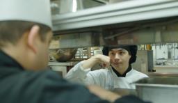 A student in a chef hat communicates with another kitchen worker